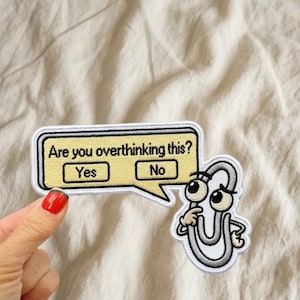 May include: An embroidered patch featuring a grey paperclip character with large eyes and a hand on its chin, next to a yellow speech bubble containing the black text Are you overthinking this? with Yes and No buttons inside the box.