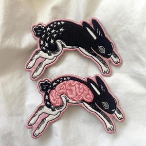 May include: Two embroidered patches shaped like leaping rabbits with black fur, white underbellies, and white star patterns. One rabbit features a visible pink brain graphic on its side, while the other shows a solid black side with white stars.