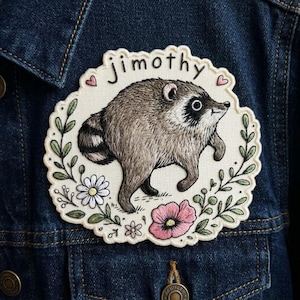 May include: An embroidered patch on a dark blue denim jacket featuring a brown and grey raccoon. The raccoon is surrounded by a floral wreath with white daisies and a pink flower. The name jimothy is stitched in black above the raccoon.