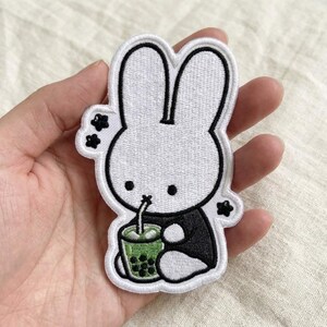 May include: An embroidered patch featuring a white rabbit character with black outlines, wearing a black shirt and drinking from a green boba tea cup with black pearls. The design includes small black star-like accents around the rabbit's head.