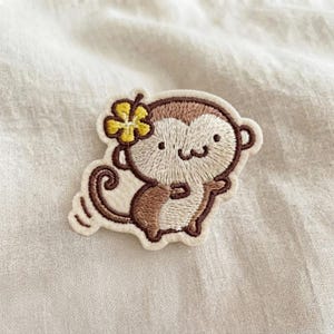 May include: An embroidered patch featuring a cartoon monkey with brown and cream-colored thread. The monkey has a small yellow flower detail on its head, a curled tail, and a simple stitched face with two black dots for eyes and a curved mouth.