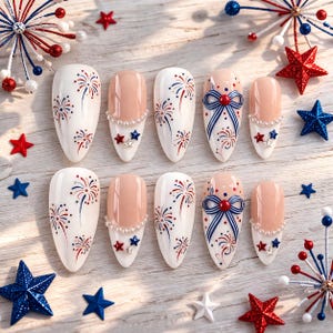 May include: A set of ten artificial nails with a patriotic theme. The nails feature white, nude, red, and blue designs, including fireworks, bows, and star accents. The nails are arranged on a light wood surface with star-shaped decorations.