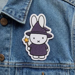 May include: An embroidered patch featuring a white rabbit character wearing a purple pointed hat and a matching purple robe. The rabbit holds a small yellow star-shaped wand in one hand. The design is outlined in white thread and has a clean finish.