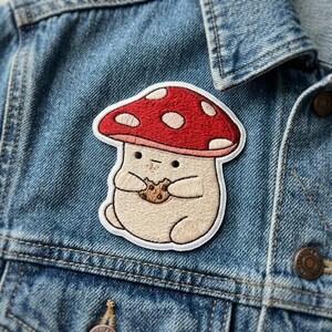May include: An embroidered patch featuring a round, beige, anthropomorphic mushroom character with a red cap adorned with white spots. The character has small black eyes and is holding a brown, textured chocolate chip cookie in its hands.