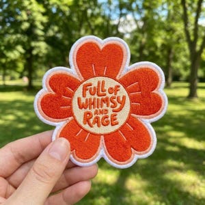 May include: A flower-shaped embroidered patch with orange petals and a cream-colored center. The text "FULL of WHIMSY AND RAGE" is stitched in orange lettering across the center. The patch features a white border around the entire shape.
