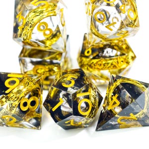 May include: A collection of clear, multi-sided dice with gold-colored numbers and internal designs. The dice are arranged in a group, showcasing their intricate details and reflective surfaces. The overall theme is gaming and tabletop accessories.