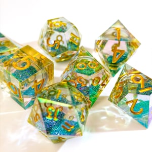 May include: A set of iridescent dice with gold numbering. The dice are clear with blue and green glitter suspended inside. The dice are various shapes, including a d20, d12, d10, d8, and d6, and are arranged on a white surface.