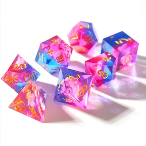 May include: A set of seven polyhedral dice in various shapes, featuring a vibrant mix of pink, blue, and purple hues. The dice have gold-colored numbers and a floral design within the translucent resin, perfect for tabletop gaming.
