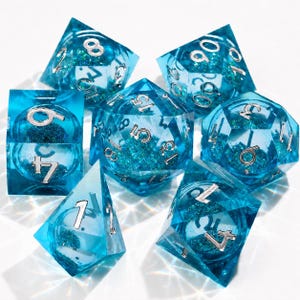 Puede incluir: Un juego de siete dados poliédricos azules transparentes con números plateados y purpurina. Los dados tienen varias formas geométricas, adecuados para juegos de rol de mesa.
