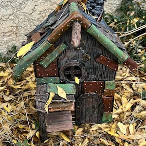 Może przedstawiać: Ręcznie wykonany domek dla wróżek o rustykalnym, teksturowanym wyglądzie. Dom posiada ciemne drewniane drzwi, okrągłe okno i komin. Dach zdobią zielone i brązowe akcenty, a konstrukcja otoczona jest opadłymi liśćmi.