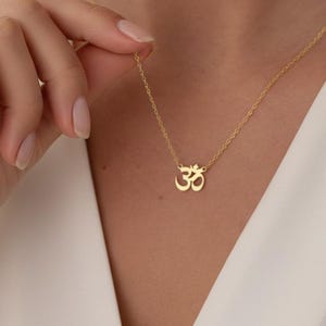 May include: A gold necklace with an Om symbol pendant. The necklace has a fine chain and a small, detailed pendant. The Om symbol is a spiritual icon, often associated with meditation and yoga. The necklace is worn around the neck.