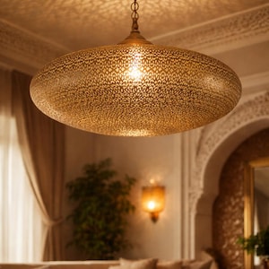 May include: A large, gold-coloured, oval-shaped pendant light with intricate cut-out patterns hangs from the ceiling. The light casts decorative shadows on the ceiling. The lamp is suspended by a chain and illuminates a living room setting.