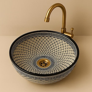 Può includere: Un lavabo rotondo decorativo con un motivo geometrico bianco e nero. Il lavabo ha uno scarico color oro ed è abbinato a un rubinetto dorato. Il design del lavabo presenta un motivo ripetuto di piccoli quadrati e punti, creando una superficie visivamente accattivante.