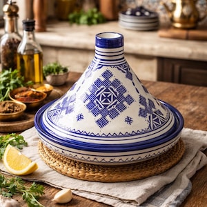 May include: A white and blue ceramic tagine pot with a conical lid, featuring geometric patterns. The tagine sits on a woven mat and a linen cloth, with ingredients like lemon and garlic nearby. A traditional cooking vessel.