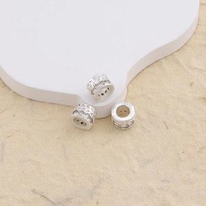 Peut inclure: Trois perles cylindriques de couleur argentée présentant une bande centrale de pierres claires et scintillantes, bordées de bords métalliques texturés en forme de tresse. Chaque perle possède un centre creux pour le passage d'un cordon.