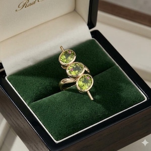 Könnte beinhalten: Ein goldener Ring mit drei ovalen, hellgrünen Edelsteinen, die jeweils mit einem kleinen rosa Stein akzentuiert sind. Der Ring wird in einer schwarzen Box mit grünem Samt präsentiert. Einzigartiges, künstlerisches Design.