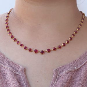 Op de afbeelding: Een goudkleurige ketting met een reeks ronde, robijnrode edelstenen. De edelstenen zijn gelijkmatig verdeeld over de ketting, wat een delicaat en elegant ontwerp creëert. De ketting wordt getoond tegen een neutrale achtergrond.