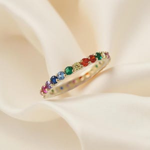 Peut inclure: Une bague dorée ornée d'une rangée continue de pierres précieuses multicolores. Les pierres présentent un arc-en-ciel de couleurs, dont le rouge, l'orange, le jaune, le vert, le bleu et le violet. La bague est posée sur un tissu blanc cassé.