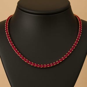 Puede incluir: Un collar rojo rubí con una cadena dorada. El collar se exhibe en un maniquí negro. El collar está hecho de pequeñas piedras redondas de color rubí. La cadena dorada es visible entre las piedras.