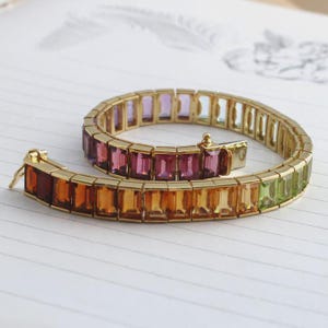 May include: A gold bracelet with a series of rectangular gemstones in a rainbow of colors, including red, orange, yellow, green, purple, and pink. The bracelet is open and lying on a white surface.
