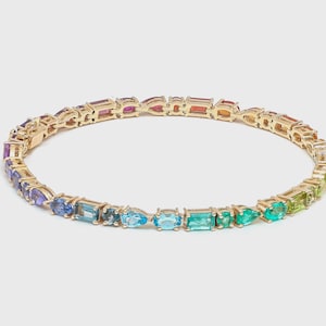 Pode incluir: Uma pulseira dourada com um espectro de pedras preciosas coloridas. As pedras estão dispostas em gradiente, passando do roxo ao verde. A pulseira está sobre um fundo branco liso.
