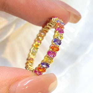 May include: A gold ring featuring a continuous row of colorful gemstones. The gemstones are oval-cut and arranged in a rainbow of colors, including pink, orange, yellow, purple, and blue. The ring is held between two fingers.