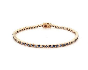 May include: A gold-toned bracelet with a row of small, round, blue gemstones. The bracelet is set against a white background. The clasp is visible and matches the gold tone of the bracelet.