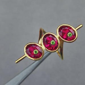 Può includere: Un anello d'oro con tre pietre preziose ovali rosso rubino, ciascuna con una piccola pietra verde al centro. L'anello ha un design unico con una fascia curva, su sfondo grigio.