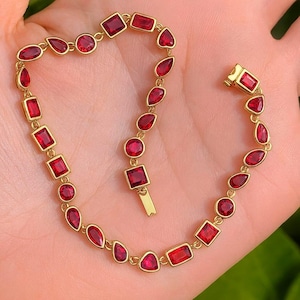 Puede incluir: Una pulsera dorada con una serie de piedras preciosas rojas de varias formas, incluyendo cuadrados, círculos y lágrimas. La pulsera tiene un cierre y parece ser de oro. Las piedras preciosas son de un color rojo vibrante.