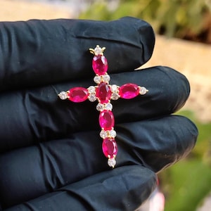 May include: A gold-coloured cross pendant with oval-cut red gemstones and small, round, clear gemstones. The cross has a crown-like design at the top. The pendant is held against a black background.