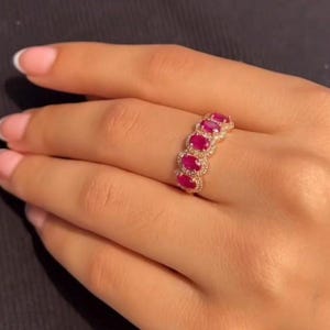 Peut inclure: Une bague en or ornée de cinq pierres précieuses rouges vives de forme ovale. Les pierres sont entourées de petits diamants transparents. La bague est portée sur une main aux ongles manucurés.