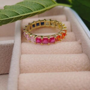 Puede incluir: Un anillo dorado con una banda de piedras preciosas cuadradas multicolores. Las piedras preciosas cambian de color, de rosa a naranja, amarillo y azul. El anillo se muestra en un joyero rosa claro, con una hoja verde en el fondo.