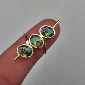 Könnte beinhalten: Ein goldener Ring mit drei ovalen, grünen Edelsteinen, jeweils mit einem kleinen roten Stein in der Mitte. Der Ring hat ein modernes und elegantes Design, wobei die Edelsteine linear angeordnet sind.