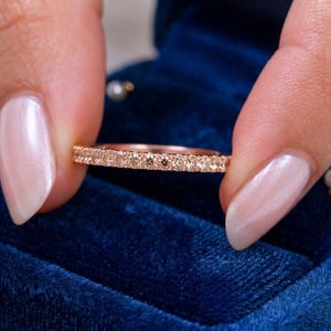 Può includere: Un anello in oro rosa con una fila continua di piccole pietre preziose scintillanti. L'anello è tenuto tra due dita su una superficie di velluto blu scuro, con piccole perle sparse.