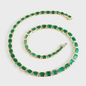 May include: A gold necklace featuring emerald-cut green gemstones. The necklace is arranged in a spiral shape, showcasing the vibrant green stones set in a gold frame. The clasp is visible at the end of the necklace.