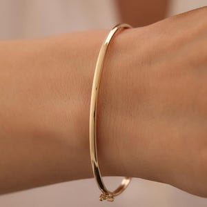 Op de afbeelding: Een gepolijste gouden armband met een glad, afgerond oppervlak en een veilige scharniersluiting. Het metaal heeft een reflecterende, hoogglanzende afwerking die het licht opvangt en een strakke, eenvoudige cirkelvormige silhouet creëert.