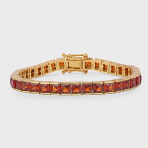 Pode incluir: Uma pulseira dourada com pedras preciosas vermelho-alaranjadas de corte quadrado. A pulseira tem um fecho seguro. As pedras são uniformemente espaçadas e engastadas numa moldura de metal dourado, criando um visual luxuoso.