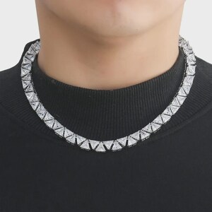 Op de afbeelding: Een zilverkleurige ketting met een reeks driehoekige, heldere edelstenen. De ketting wordt gedragen over een zwarte coltrui. De edelstenen zijn dicht op elkaar geplaatst, waardoor een doorlopende, sprankelende band om de hals ontstaat.