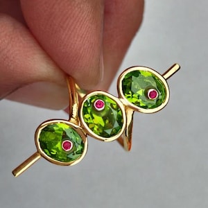 Op de afbeelding: Een gouden ring met drie ovale groene edelstenen, elk met een kleine rode accent. Het ontwerp van de ring is modern en elegant, met een minimalistische uitstraling. De edelstenen zijn gezet in een gouden band.