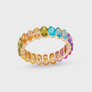 Puede incluir: Un anillo dorado con una fila continua de piedras preciosas de corte ovalado en un arcoíris de colores. Las piedras preciosas incluyen tonos de naranja, amarillo, verde, azul y morado. El anillo está sobre un fondo neutro.