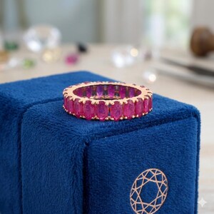 May include: A gold-toned eternity ring set with oval-cut, vibrant pink gemstones. The ring is displayed on a dark blue velvet surface, showcasing the ring's color and design. The gemstones are closely set, creating a continuous band of color.