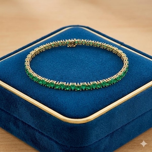 May include: A gold-toned bracelet with a row of emerald-green gemstones. The bracelet is displayed on a blue velvet jewelry box with gold trim. The gemstones are evenly spaced and create a continuous line around the wrist.