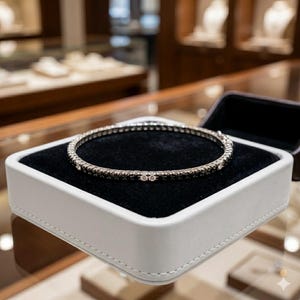 May include: A silver bracelet with small, sparkling stones is presented in a white jewellery box with a black velvet interior. The circular bracelet appears to be made of metal, with the stones set closely together. The box is open, displaying the bracelet.