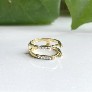 Peut inclure: Une bague dorée avec un design à double bande. Chaque bande est ornée de petites pierres précieuses claires. La bague est posée sur une surface blanche avec un arrière-plan vert flou, suggérant un thème de bijoux ou d'accessoires.