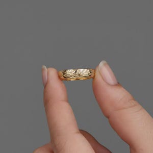 以下が含まれることがあります： 花柄のゴールドリング。リングは、灰色の背景に対して2本の指の間に保持されています。リングの表面は、葉と花の精巧な彫刻でテクスチャ加工されています。