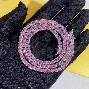 以下が含まれることがあります： 小さな正方形のピンク色の宝石が連なった、きらびやかなネックレス。複数のストランドに配置され、重ね着のような効果を生み出しています。留め金はシルバーカラーです。黒い背景に展示されています。