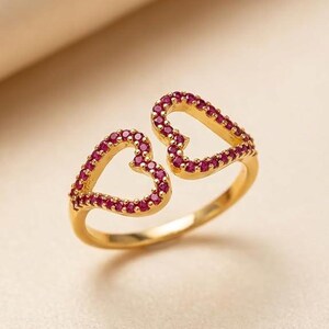Könnte beinhalten: Ein goldfarbener Ring mit zwei offenen Herzen. Jedes Herz ist mit kleinen, roten Edelsteinen besetzt. Der Ring ist vor einem weichen, neutralen Hintergrund platziert, um das Design und die Farbe des Schmucks hervorzuheben.