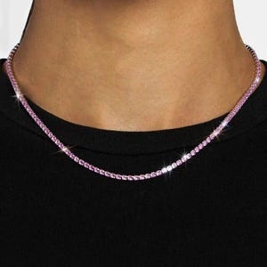 May include: A pink tennis necklace with small, sparkling gemstones. The necklace is worn against a black top, highlighting the vibrant colour and intricate design of the jewellery. The necklace appears to be made of metal.