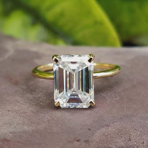 May include: A gold ring featuring a large, rectangular, clear gemstone. The gemstone is set with four prongs and has a step-cut design. The ring band is a simple, polished gold. The background is a blurred green and brown.