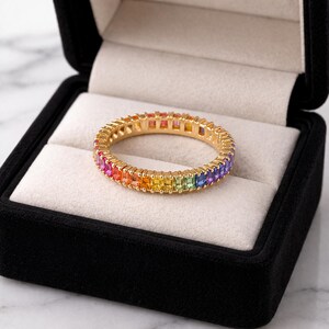 Puede incluir: Un anillo dorado con una banda completa de piedras preciosas rectangulares multicolores. Las piedras están dispuestas en un patrón de arcoíris, pasando del rojo al morado. El anillo se muestra en una caja de joyería negra con interior blanco.